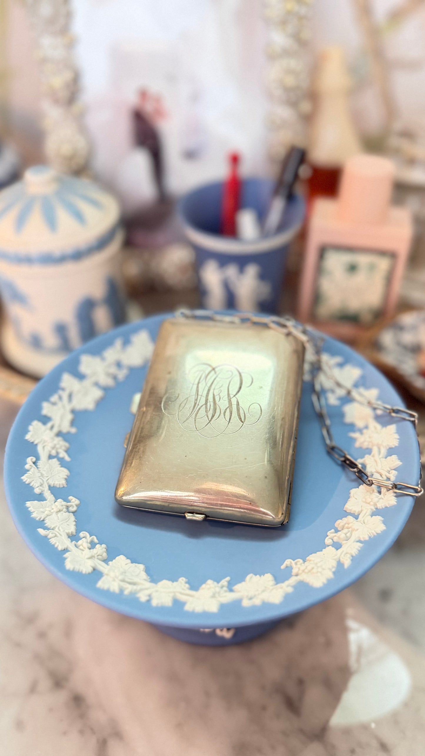 Antique German-silver dance purse with engraved script monogram and chain displayed on a blue Wedgwood pedestal.