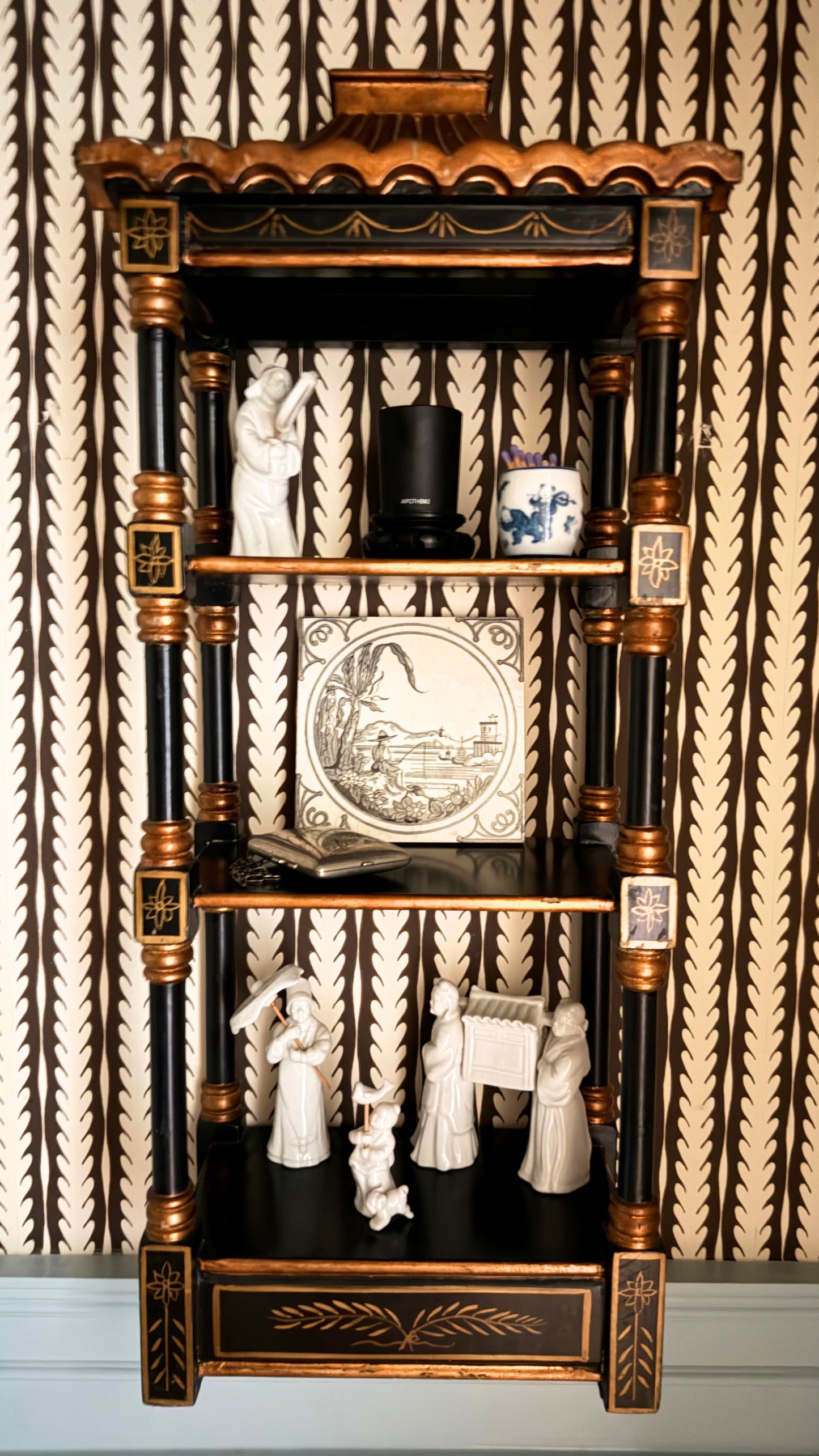Vintage black and gold chinoiserie pagoda wall shelf styled with porcelain, a Minton tile, and silver purse against brown Oceana wallpaper.